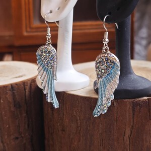 May include: A pair of silver-toned earrings shaped like angel wings. The wings feature a light blue enamel and are embellished with clear rhinestones. The earrings are displayed on white and black earring stands against a wooden background.