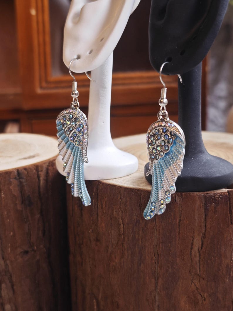 May include: A pair of silver-toned earrings shaped like angel wings. The wings feature a light blue enamel and are embellished with clear rhinestones. The earrings are displayed on white and black earring stands against a wooden background.