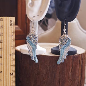 May include: A pair of silver-toned wing earrings with blue and white enamel detailing and sparkling crystal accents. The earrings are displayed on ear-shaped stands, showcasing their design. The earrings are approximately 2 inches in length.