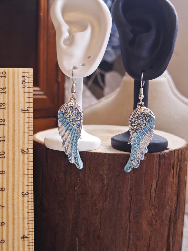 May include: A pair of silver-toned wing earrings with blue and white enamel detailing and sparkling crystal accents. The earrings are displayed on ear-shaped stands, showcasing their design. The earrings are approximately 2 inches in length.
