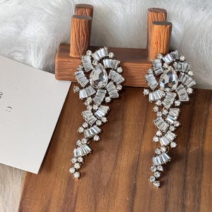 Puede incluir: Un par de pendientes de cristal brillantes. Cada pendiente presenta una gran piedra central en forma de lágrima, rodeada de cristales rectangulares más pequeños. Una cascada de cristales cuelga debajo, creando un diseño glamuroso.