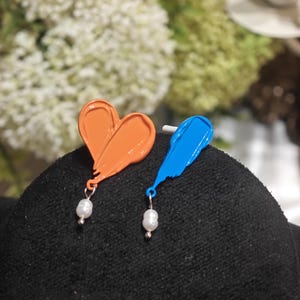 May include: A pair of earrings, one orange heart-shaped and the other blue feather-shaped, each with a small white pearl. The earrings are set against a dark background, with blurred greenery in the background.