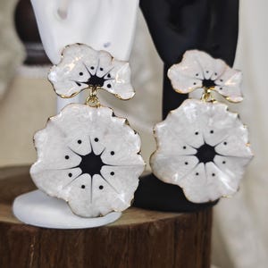 May include: A pair of white flower earrings with a black centre and gold edging. The earrings are displayed on white and black earring stands. The earrings have a layered design, with a smaller flower shape at the top and a larger one below.