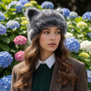 May include: A gray faux fur hat with cat ears. The hat is worn by a person wearing a white collared shirt, a green sweater, and a brown blazer. The background features a variety of colorful hydrangea flowers.
