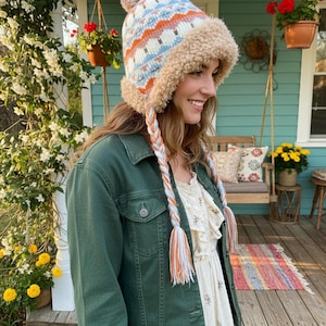 May include: A patterned winter hat with a pom-pom, in white, blue, and orange. It has a faux fur trim and braided ear flaps with tassels. This hat is part of a winter clothing collection.