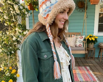 May include: A winter hat with a pom-pom, featuring a pattern of white, blue, and orange. The hat has a faux fur trim and braided ear flaps with tassels. The hat is part of a winter clothing collection.