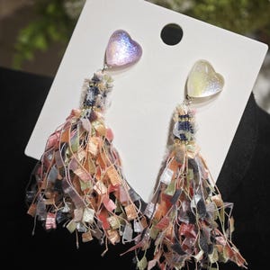 Può includere: Un paio di orecchini pendenti con top a forma di cuore e frange di nappe colorate. Gli orecchini sono attaccati a una carta bianca. Le nappe sono di vari colori tra cui rosa, arancione e verde. I top a forma di cuore hanno glitter iridescenti.