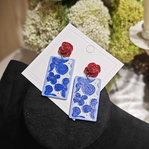 May include: A pair of earrings featuring a rectangular design with blue floral patterns on a white background. Each earring has a small red rose at the top. The earrings are displayed on a white card.