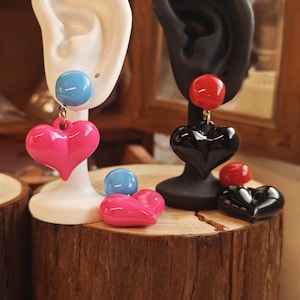 May include: Earrings with heart and round bead designs. One pair features a pink heart and blue bead, displayed on a white ear model. The other pair has a black heart and red bead, shown on a black ear model. Additional loose beads and hearts are also present.