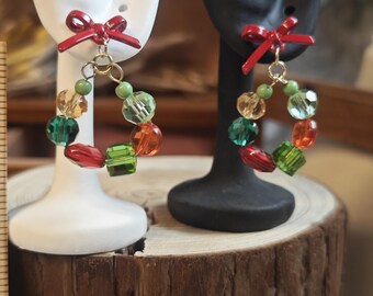 May include: A pair of hoop earrings featuring a red bow detail. The hoops are decorated with a variety of colourful beads in green, orange, and clear. The earrings are displayed on white and black earring stands.