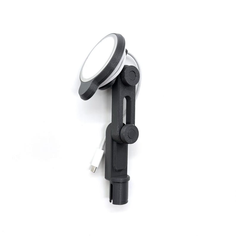 May include: A dark gray adjustable mounting arm featuring a circular magnetic charging pad at the top. The device includes two rotating joints with circular knobs for positioning and a white USB-C charging cable attached to the side.