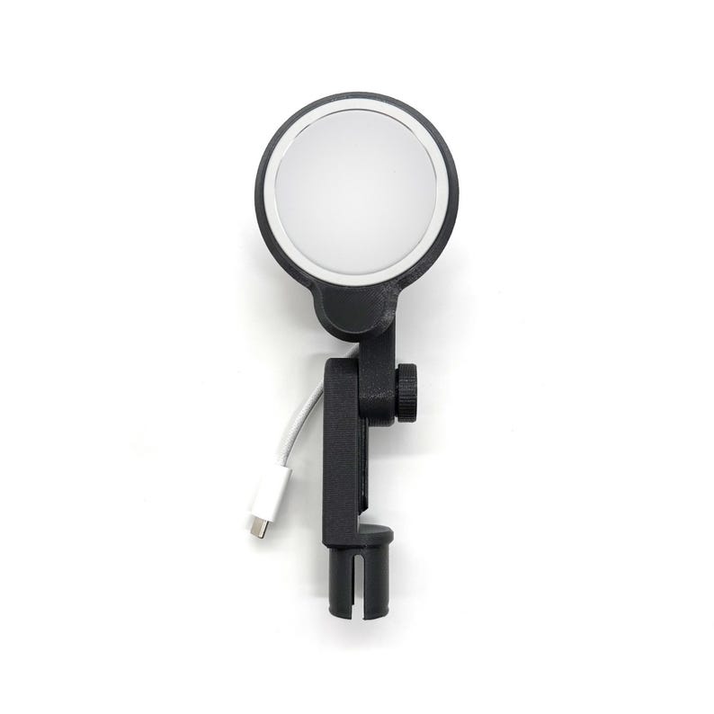 May include: A black LED ring light with a white circular light and a black adjustable clamp. The light has a white USB-C cable attached. The ring light is designed for use with a phone or camera.