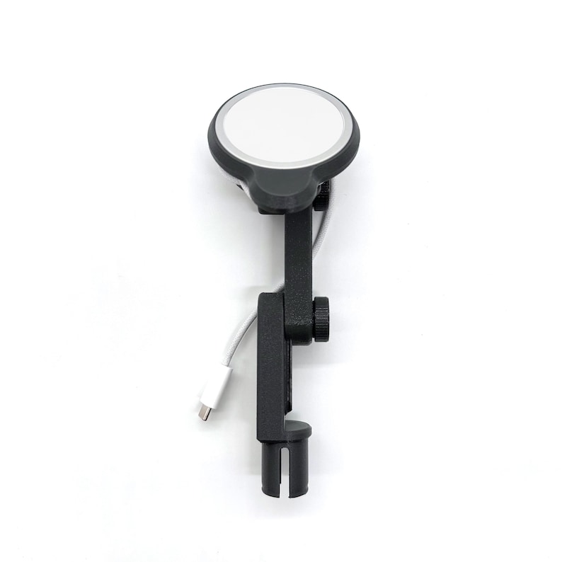 May include: A black adjustable phone holder with a white circular charging pad. The holder has a clamp for securing to a surface and a USB-C cable. The charging pad has a white surface and a black border.