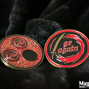 Puede incluir: Dos pines circulares de metal esmaltado con un esquema de color negro y rojo. Un pin presenta tres símbolos de espiral giratorios rodeados de patrones de llamas. El otro pin muestra un gráfico de espada y el texto rojo go again en diseño.
