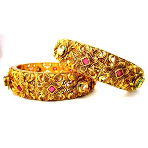 May include: Two gold bangles with intricate floral designs and pink and green gemstones.