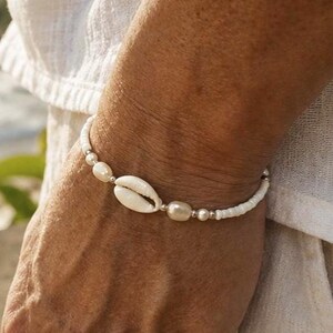 May include: A beaded bracelet featuring a central white cowrie shell, flanked by smooth off-white pearls and small silver-toned metallic spacer beads, strung together with a series of tiny white seed beads completing the circular band design.