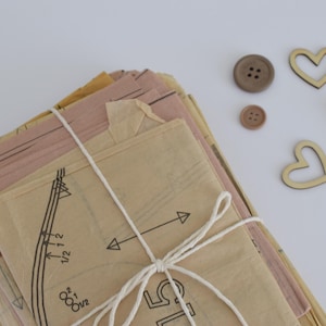 May include: A bundle of vintage sewing patterns tied with white twine, alongside wooden buttons, heart-shaped cutouts, and a small clothespin. The patterns are in shades of beige and pink.