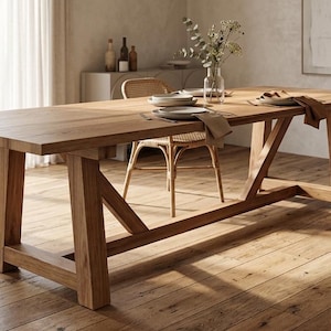 May include: A long rectangular wooden dining table with a trestle base featuring angled support beams. The table is accompanied by several light brown woven rattan chairs with curved armrests and backrests, arranged around the tabletop.