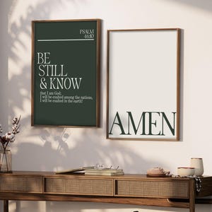 Könnte beinhalten: Zwei gerahmte Kunstdrucke mit religiösem Text. Einer zeigt "BE STILL & KNOW" mit einem Bibelvers, der andere sagt "AMEN". Der Text ist dunkelgrün und weiß auf neutralem Hintergrund, in Holz gerahmt. Sie hängen über einer Holzkonsolentisch.