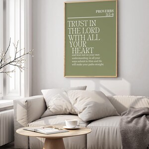 May include: A framed print with the text "TRUST IN THE LORD WITH ALL YOUR HEART" in white on a green background. The print is hanging above a light-colored sofa with pillows and a coffee table with a cup and saucer.