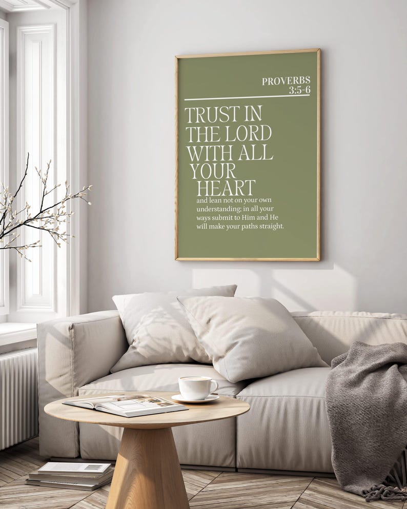 May include: A framed print with the text "TRUST IN THE LORD WITH ALL YOUR HEART" in white on a green background. The print is hanging above a light-colored sofa with pillows and a coffee table with a cup and saucer.