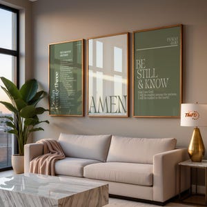 May include: Three framed wall art pieces with gold frames. The art features text in white on a green background. The middle frame says "AMEN". The right frame says "BE STILL & KNOW". A beige sofa and marble coffee table are in front of the art.