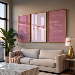 Op de afbeelding: Drie ingelijste wandkunstwerken met gouden frames en roze achtergronden. De kunst bevat tekst, waaronder "Romeinen 8:28", "PURPOSE" en "Jeremia 29:11". Een beige bank, een marmeren salontafel en een gouden lamp staan op de voorgrond.