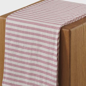 May include: A pink and white striped table runner draped over a wooden surface. The runner features horizontal stripes and is made of a soft, textured fabric. The wooden surface has a natural wood grain.