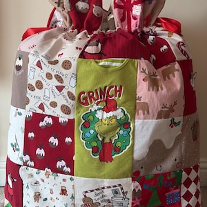 May include: A large, rectangular fabric sack constructed from various Christmas-themed patches. The centre features a green panel with a Grinch graphic and the text 'GRINCH'. Other patches display gingerbread men, reindeer, puddings, and text reading 'Santa is coming' and 'Dear Santa'. The top is gathered with a red ribbon.