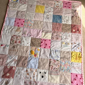 May include: A patchwork memory quilt constructed from various baby clothing fabrics in shades of pink, white, yellow, and tan. The quilt features a variety of patterns including hearts, florals, animal prints, and text such as Mummy, Daddy, and Easter.