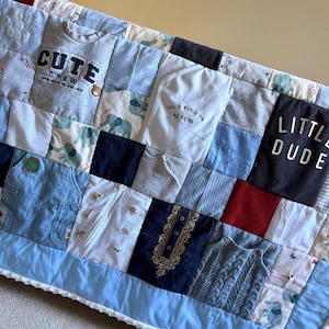 May include: A baby quilt made from repurposed baby clothes. The quilt is a patchwork of various fabrics, including denim and knitwear. Some squares feature text such as "CUTE CREW", "HELLO WORLD I'M ADYAAN", and "LITTLE DUDE". The quilt is trimmed with a light blue border.