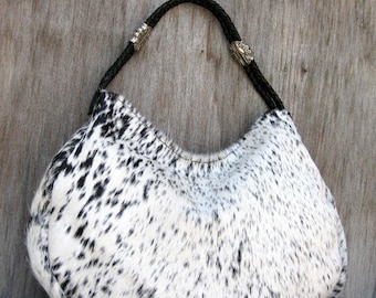 May include: A cowhide hobo bag with a black braided handle and silver-toned metal accents. The bag features a white background with black spotted markings, and a curved, crescent shape.