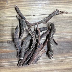 May include: A collection of dried, dark brown roots and stems, possibly herbs or spices, arranged on a light brown wooden surface. The pieces vary in size and shape, with some appearing rough and textured.