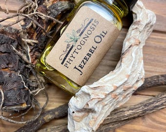 May include: A small glass bottle of "Jezebel Oil" with a black cap and a brown label featuring the text "Phytognosis". The bottle is surrounded by dried plants and wood pieces, all set on a wooden surface.