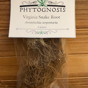 May include: A clear plastic bag containing dried Virginia Snake Root, labelled with the text "PHYTOGNOSIS" and the botanical name "Aristolochia serpentaria." The label also indicates a weight of 5 grams. The background is a wooden surface.