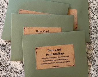May include: Three green envelopes with brown labels that read "Three Card Tarot Readings". The label explains that inside each envelope are three tarot cards that represent past challenges, current areas of focus, and future prospects.