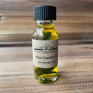 May include: A small glass bottle with a black cap filled with a yellow liquid and green leaves. The label reads "PHYTOGNOSIS Abre Camino Oil www.phytognosis.org"