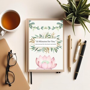 以下が含まれることがあります： 「10 Minutes for You.」の文字とピンクの蓮の花のイラストが入ったベージュ色のジャーナル。ジャーナルの周りにはペン、お茶のカップ、メガネ、鉢植えの植物があります。ジャーナルは心と精神のためのミニジャーナルです。