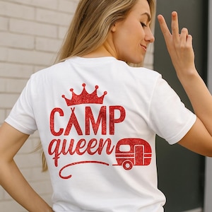 Pode incluir: Camiseta branca com um design vermelho brilhante com uma coroa, as palavras "CAMP queen" e um gráfico de trailer de campista. A camisa é feita de um material macio.