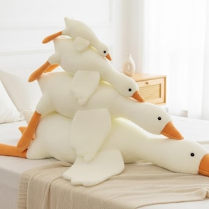Könnte beinhalten: Drei weiße Plüschgänse übereinander gestapelt. Die Gänse haben orangefarbene Schnäbel und Füße sowie schwarze Augen. Sie liegen auf einem Bett mit einer beigen Decke.