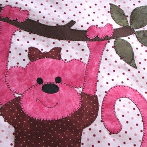 May include: A pink monkey with a brown bow hanging from a brown branch with green leaves. The monkey is on a white background with pink polka dots.