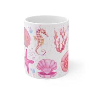 Könnte beinhalten: Weiße Keramiktasse mit einem Aquarelldesign von Meereslebewesen. Das Design umfasst ein Seepferdchen, Muscheln, Seesterne, Korallen und Blasen in Rosa-, Pfirsich- und Koralltönen. Die Tasse hat eine glänzende Oberfläche.