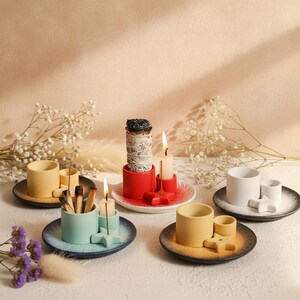 Può includere: Quattro set di portaincenso e portacandele in ceramica, ciascuno composto da un vassoio circolare, una tazza cilindrica e un piccolo supporto a forma di stella. I set sono gialli, rossi, azzurri e bianchi, con candele accese e bastoncini d'incenso.
