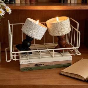 May include: Two lit candles with floral designs in a white wire basket. One candle is white, the other is beige. The candles are on black and brown stands. The basket rests on books with the titles "Full of Luck", "Butter", and "Around".