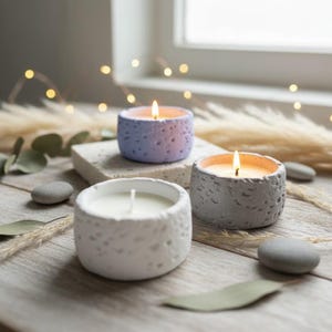 May include: Three lit candles in textured, round concrete holders. One is white, one is light purple, and one is grey. The candles sit on a wooden surface with decorative stones and dried plants, creating a cosy, natural aesthetic.