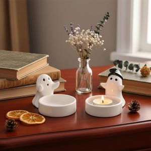 May include: White ceramic ghost-shaped candle holders with round bases. One ghost wears a black hat. A small glass vase holds dried flowers. A lit candle sits in one holder. Decorative items on a wooden table.
