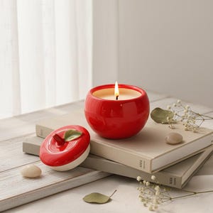 Puede incluir: Una vela roja de cerámica con forma de manzana, con una mecha encendida. La vela está sobre una pila de libros, con la tapa en forma de manzana al lado. La tapa tiene un detalle de hoja verde. También hay piedras decorativas y ramitas de flores blancas.