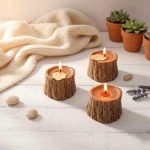 May include: Three brown tree stump-shaped candles with lit wicks. The candles sit on a white wooden surface, with a cream-colored blanket in the background. Small potted succulents and decorative stones are also present.