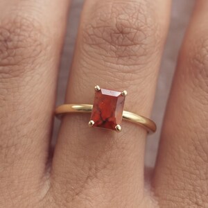 May include: A gold ring featuring a rectangular, emerald-cut orange stone with dark red veining. The stone is held in a four-prong setting on a smooth, thin gold band. The design is simple and focuses on the contrast between the metal and stone.