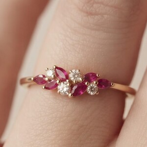 May include: A gold ring featuring a cluster of marquise-cut red rubies and round-cut white diamonds. The stones are arranged in a delicate, asymmetrical floral-inspired pattern along the thin, polished gold band, creating a refined look.